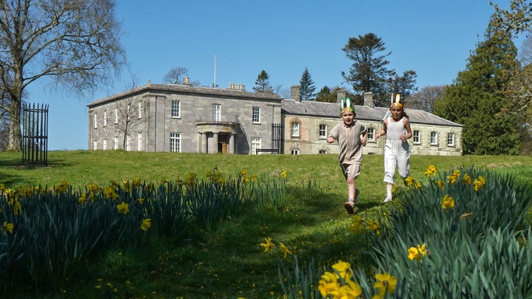 Two children running on a path through the daffodils with a mansion behind them
